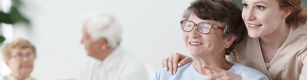 Banner picture of a female Nurse standing behind an elderly woman and pointing. There is a faded background of an elderly woman and man looking at each other.