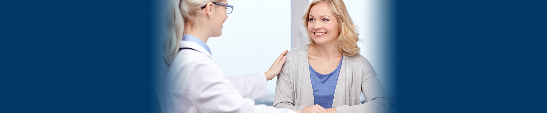 Banner picture of a female physician looking at a female patient. She has her hand on her shoulder and they are both smiling.