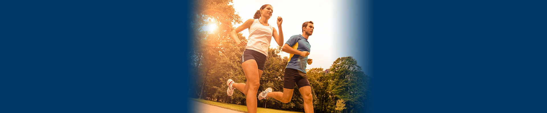Banner picture of a female and male outside jogging. There is the sun beaming through the trees behind them as they run.