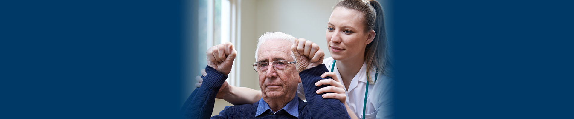 Banner picture of a elderly male patient doing Neurological Rehab. There is a female Medical Professional behind him lifting his arms up as he clenches his fist.