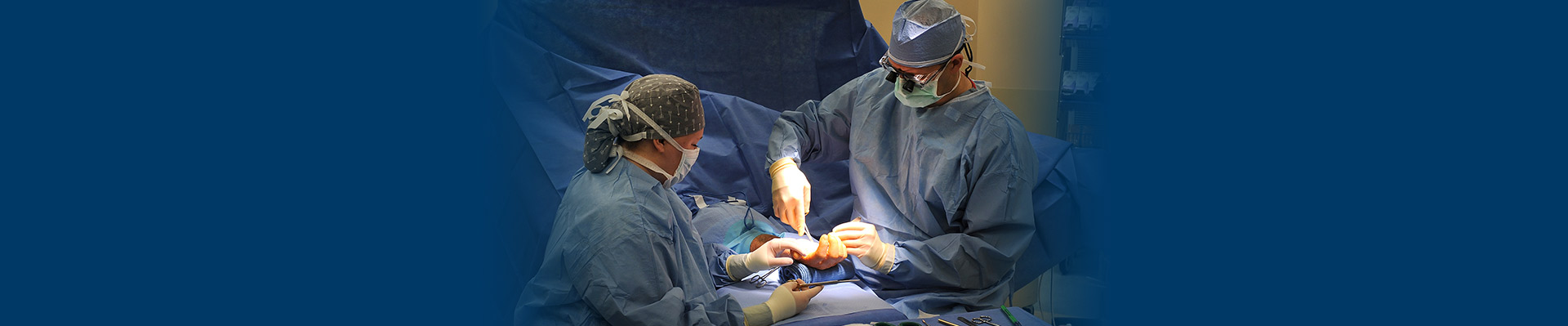 Banner picture of a male surgeon and a female surgeon about to perform surgery on a patients hand in an operation room.