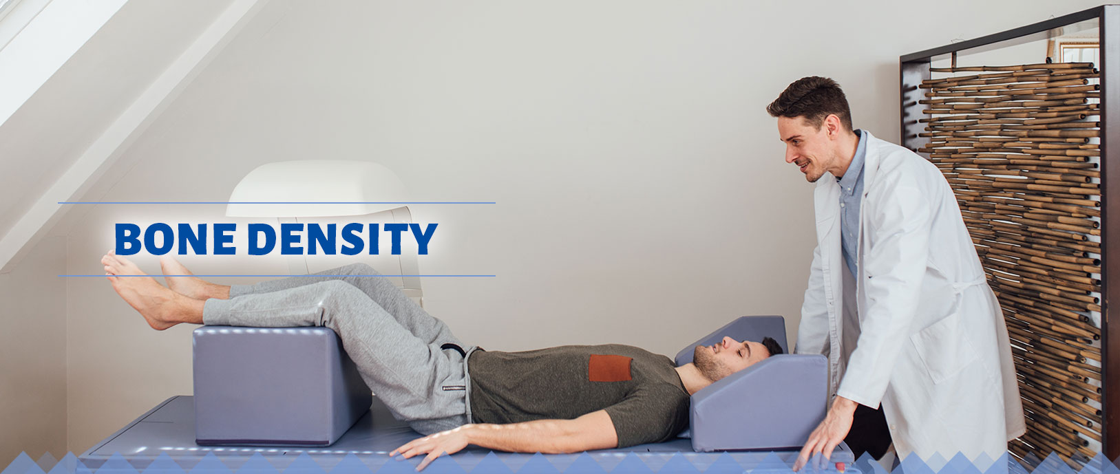 Bone density: a patient is lying on a flat surface with his legs elevated and propped onto a cushion. A bone density machine is focused above his legs and is being observed by ta smiling physician, who is at the head of the patient.