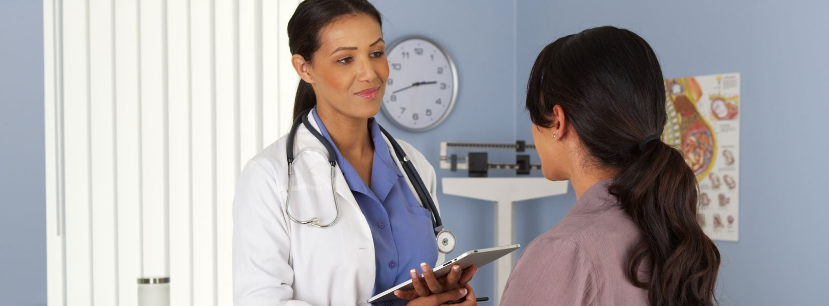 Banner picture of a female Physician standing in front of a female patient in a Doctor's office. She is wearing a medical coat , stethoscope around her neck, and holding a tablet.