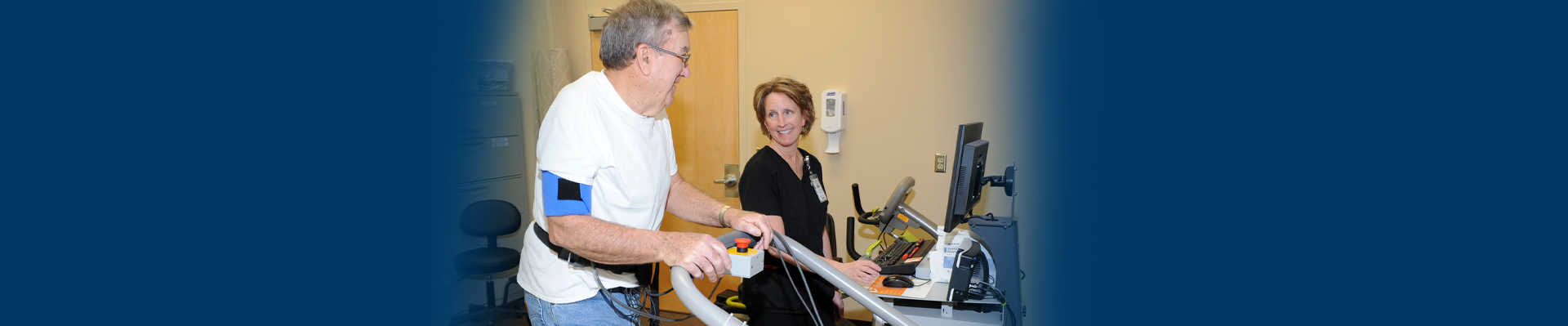 Banner picture of an elderly male patient standing up with wires hooked around his waist as he is doing Cardiac Rehab. There is a female Nurse standing next to him in front of a keyboard and computer monitor. She is smiling at the patient,