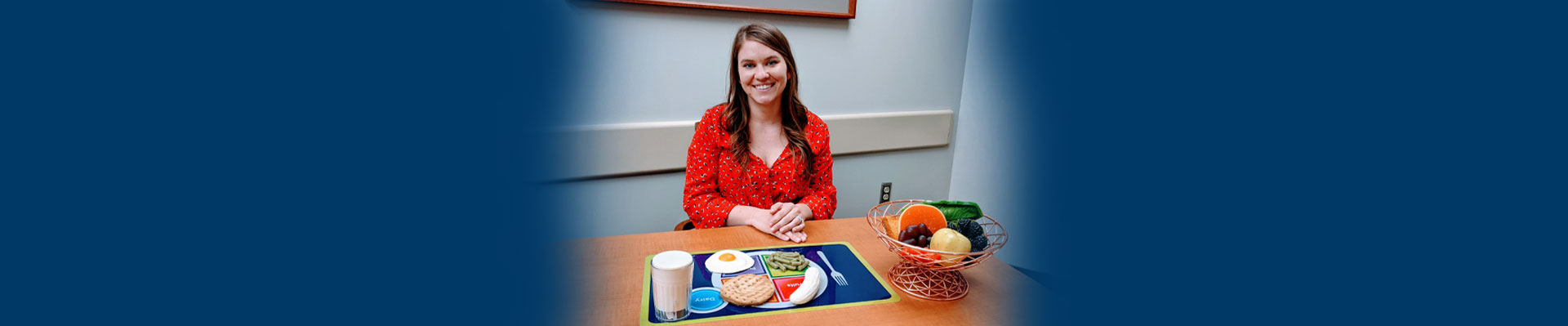 Banner picture of a female Dietician  sitting at a table with a placement mat with a plate prin with milk, an egg, green beans, a banana, bread and a basket sitting next to her full of fruit.
Dietitian Services