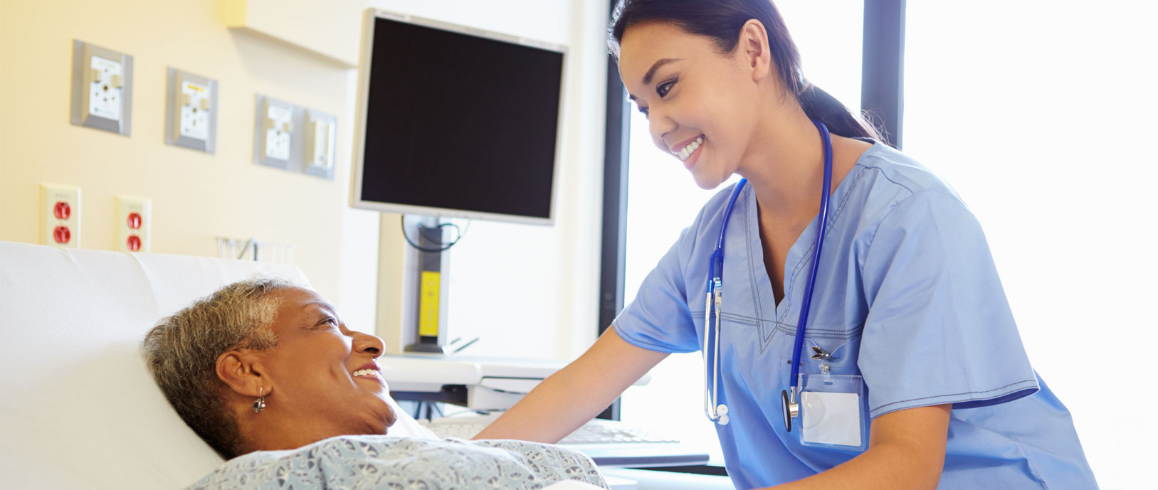 Banner picture of a smiling female Nurse standing over a smiling female patient laying in a hospital bed with a medical monitor in the background.