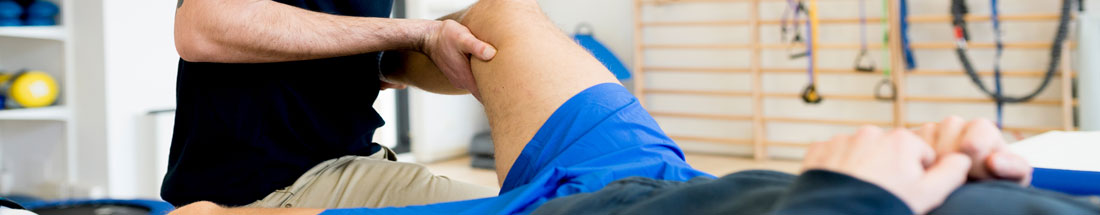 Banner picture of a male physical therapist moving a male patient leg around while the male patient is lying on his back on top of a patients table.