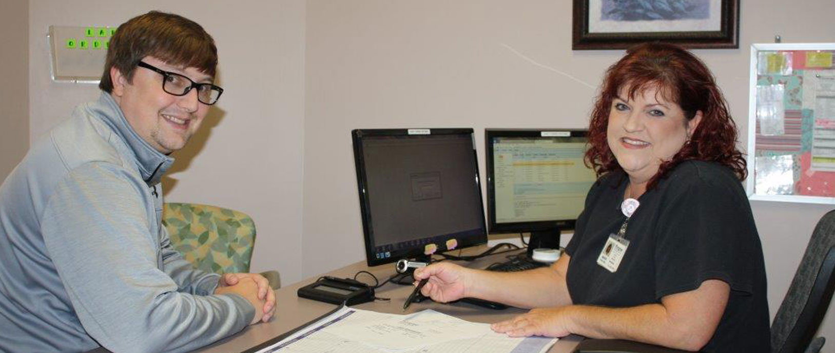 Banner picture of a female Medical Staff sitting down with a male patient at a desk in front of two computer monitors. She is holding a pen and pointing to some information on paperwork.