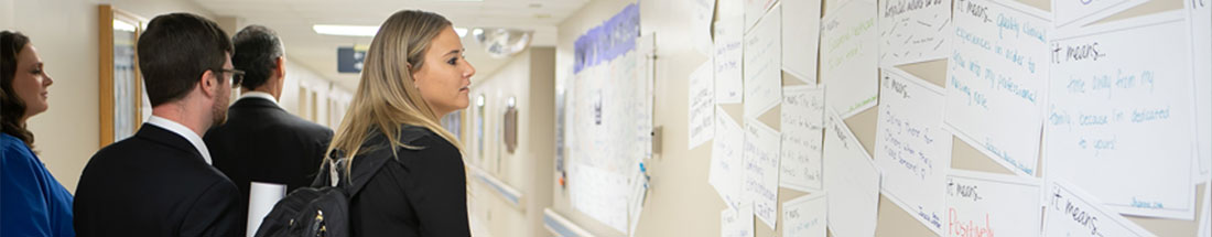 Banner picture of two females and two males dressed nicely walking down the Hospital hallway. A woman is looking at notes on a bulletin board.