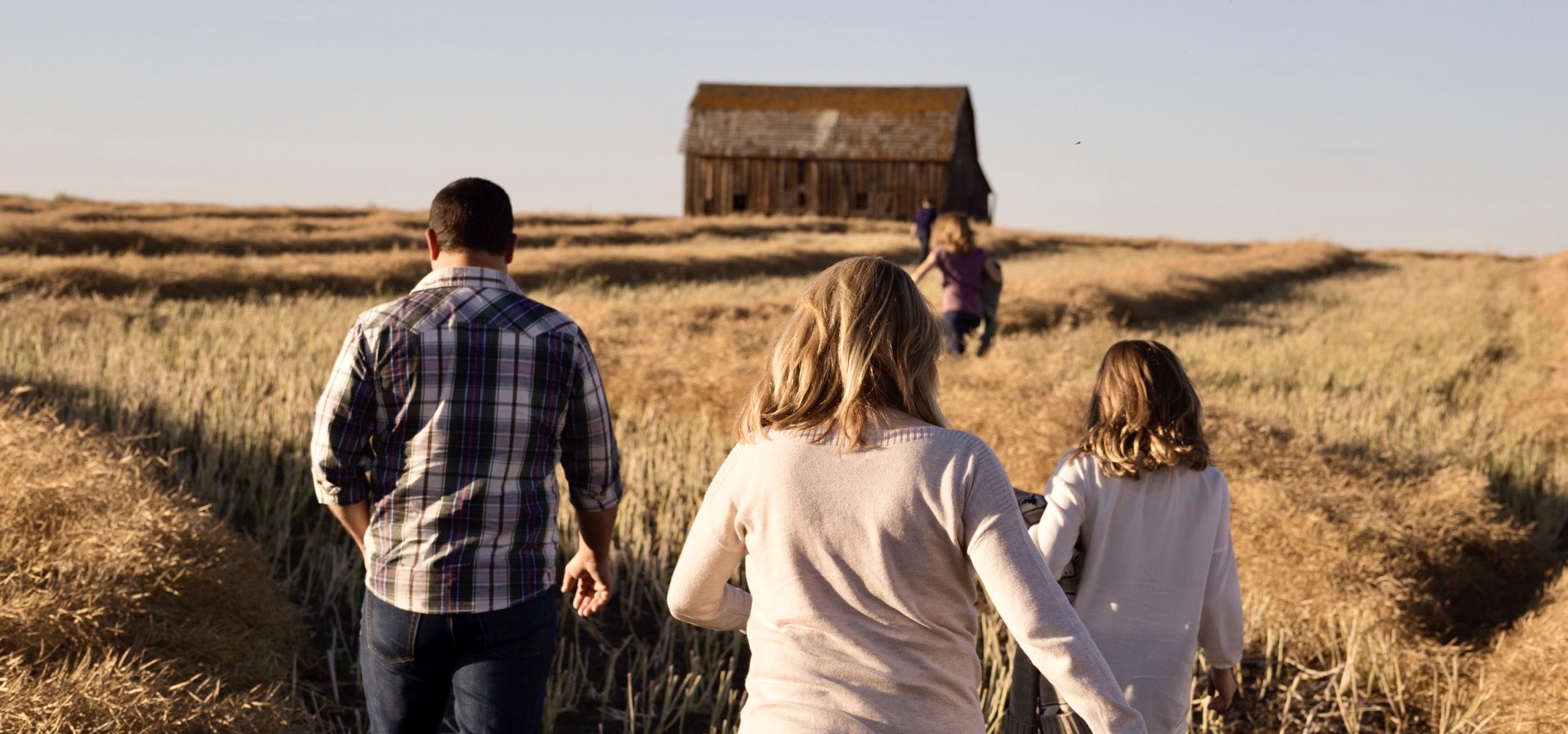Banner picture of a man, woman, and young girl walking in a grassland field.There is a p;d deserted barn out in the field.