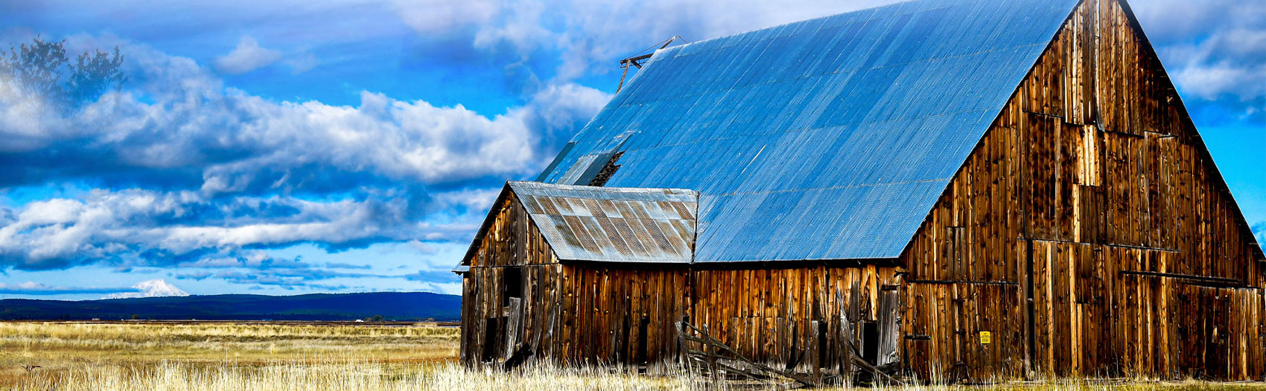 Banner picture of a large triangle shaped wooded barn on a large piece of land with some hills/mountains in the background.