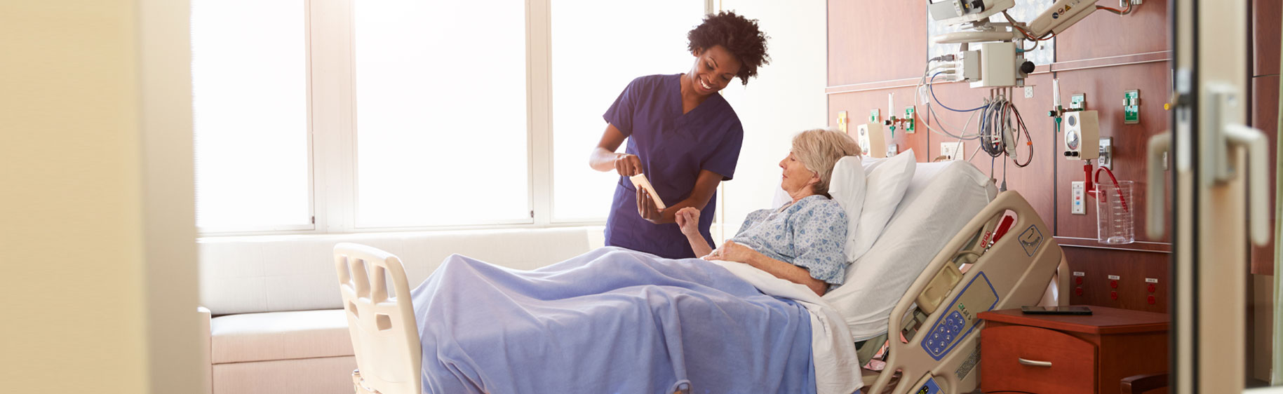 Banner picture of a female Nurse talking to an older female patient who is sitting up in a swing bed in a patient hospital room.