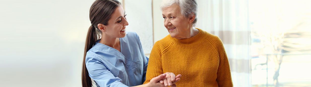 Banner picture of a young female Nurse holding an elderly woman's hand that is standing up. They are both smiling at each other.