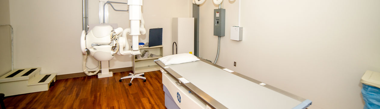 Banner picture of an X-ray machine in a room with a bed table for patients.