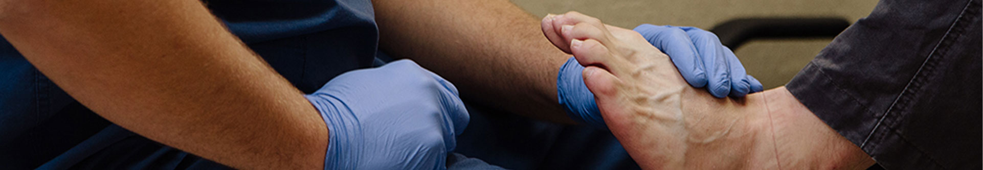 Banner picture of a male Nurse holding a patients bare foot on his lap. He is wearing gloves.