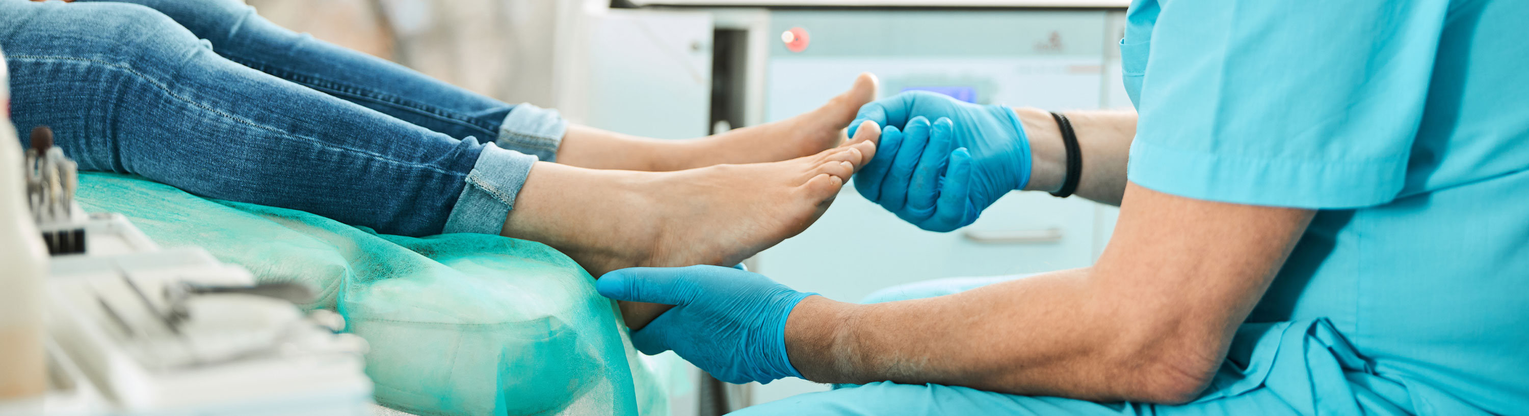 Picture of a male Podiatrist sitting down and wearing gloves while he holds a female patients heel with one hand and the other putting pressure of her big toe