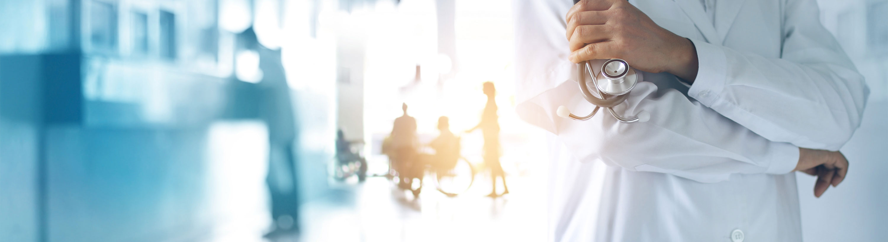 Banner picture of a male Physician standing in a hospital lobby and holding a stethoscope in one hand. There is a blurred background of a woman pushing a man in a wheelchair and a man standing up at the front desk counter.