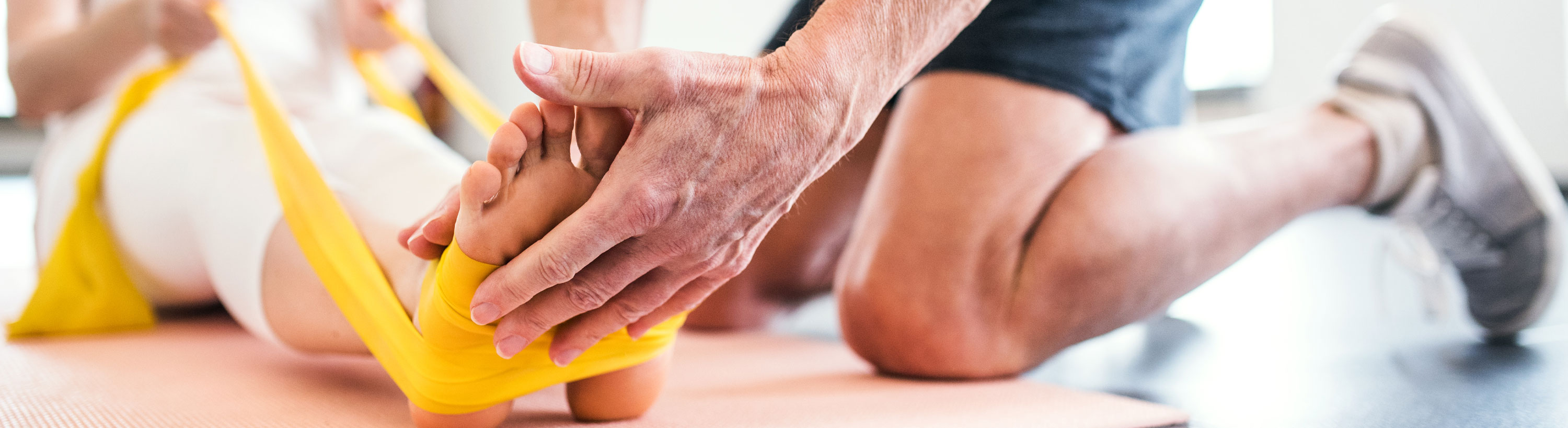 Banner Picture of a Physical Therapist on his knees bent over holding a female patients  feet on both sides as she pulls her feet up with her arms by holding onto elastic material rope