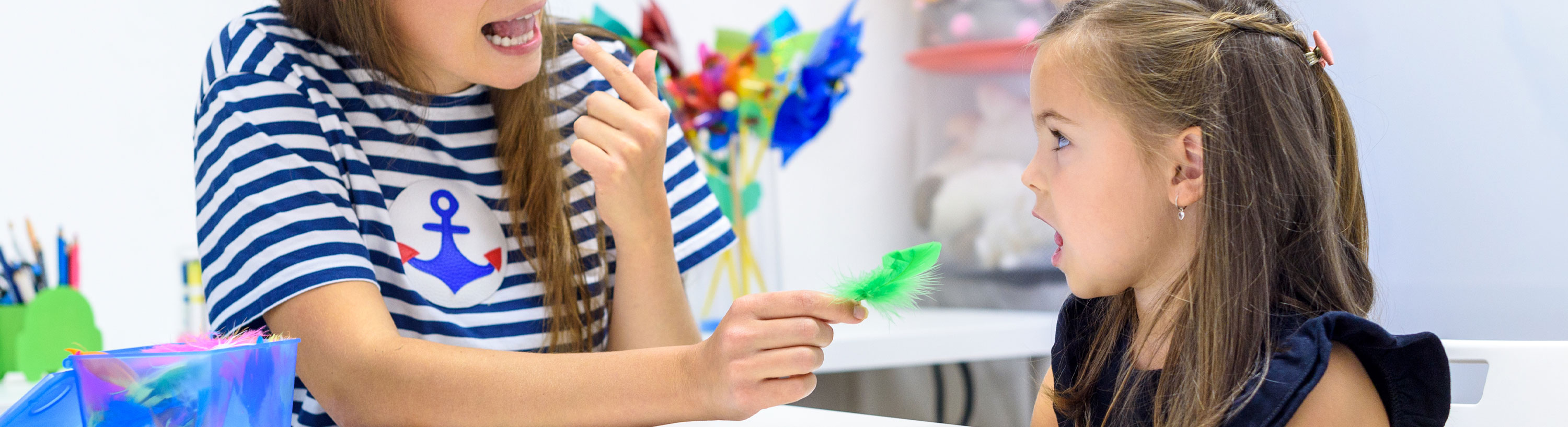 Banner picture of a female Speech Therapist holding her finger up and pointing to her tongue while rolling it as a little girl is looking at her trying to roll her tongue as well
