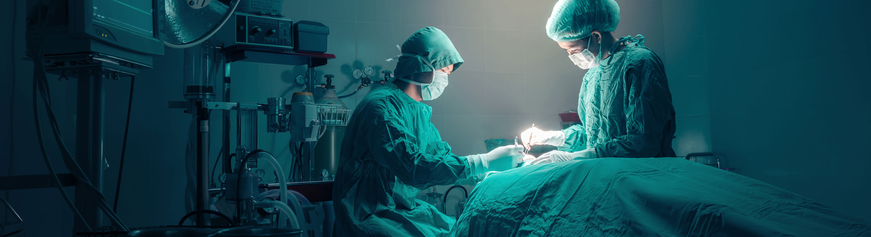 Banner picture of two surgeon's wearing medical covers over their clothes, gloves, mask, and head cap on. They are in an operation room performing surgery on a patient.