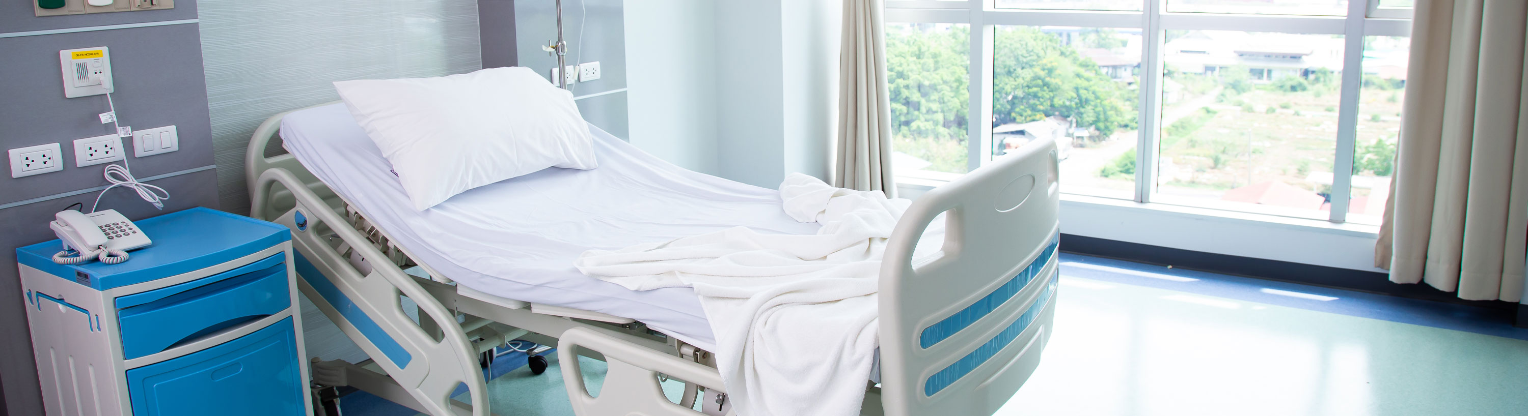 Banner picture of a patient room with a swing bed, side table, phone, IV drip roller, and pretty view outside a large indoor window.