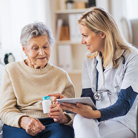 Picture of a female Nurse sitting next to an elderly female patient. The patient is holding her prescription and the nurse is pointing at it smiling.