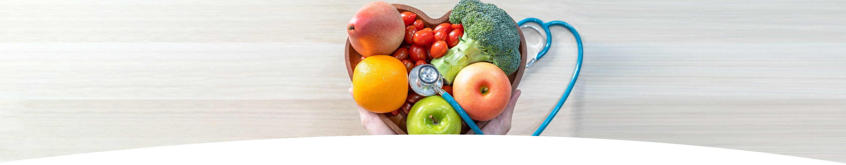 Banner picture of a heart shaped wooden bowl with cherry tomatoes, broccoli, apples, and an orange. There is a stethoscope in the bowl of the cherry tomatoes and the other end is on the table.