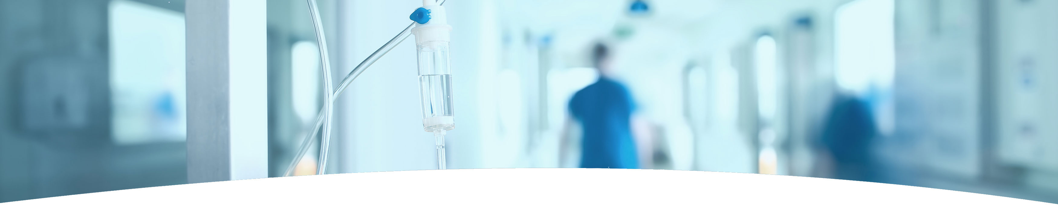 Banner picture of an IV drip that is sitting in a hospital hallway. There is a blurred background of hospital hallway and a male Nurse pushing someone in a wheelchair.