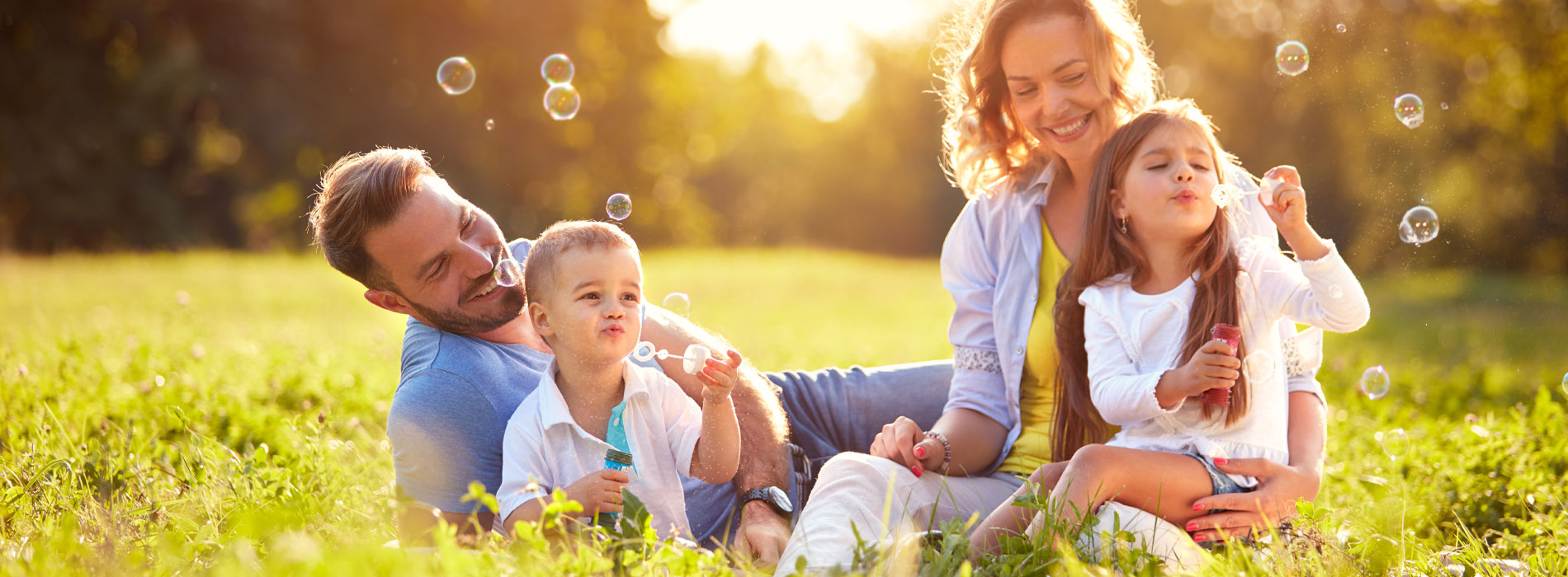 Banner picture of a couple with their son and daughter sitting and lying down in a grass field. The children are blowing bubbles and their parents are smiling.