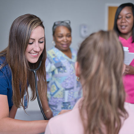 Picture of a young female Nurse using a stethoscope on a female patient. There is two female Nurses in the background smiling.
