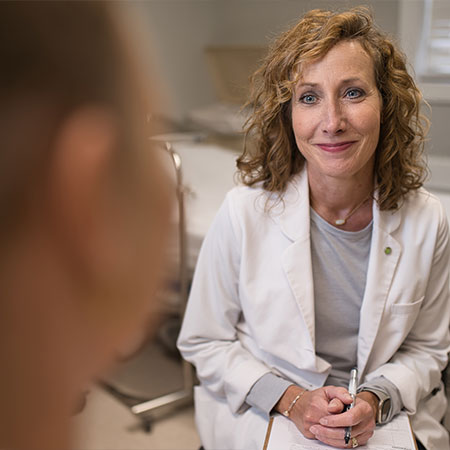 Picture of a female Physician wearing a Medical coat and sitting down holding a clipboard and pen. She is smiling at a patient who is sitting in front of her.